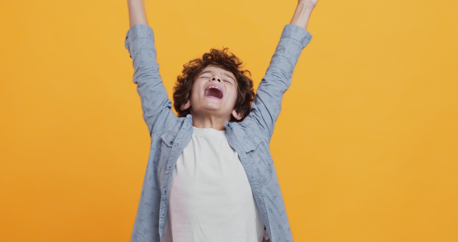 Child enjoyment. Happy boy enjoying win, screaming with raised hands, orange studio background | Shutterstock HD Video #1061034958