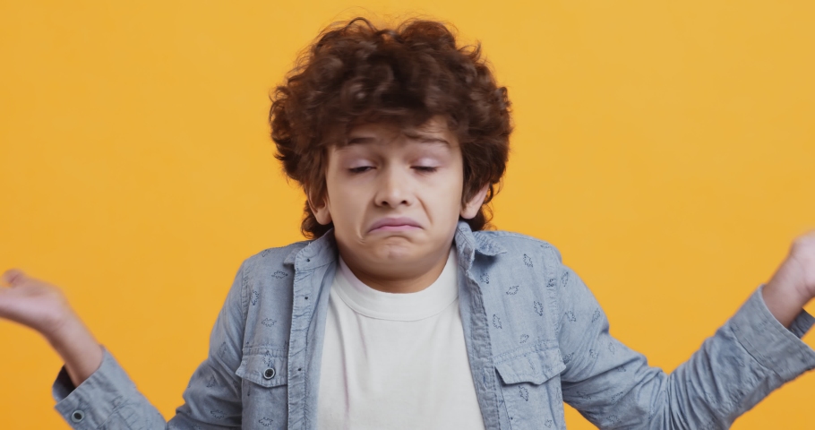 Studio portrait of unsure little curly boy shrugging shoulders and shaking head no, doubt concept, orange studio background Royalty-Free Stock Footage #1061035096