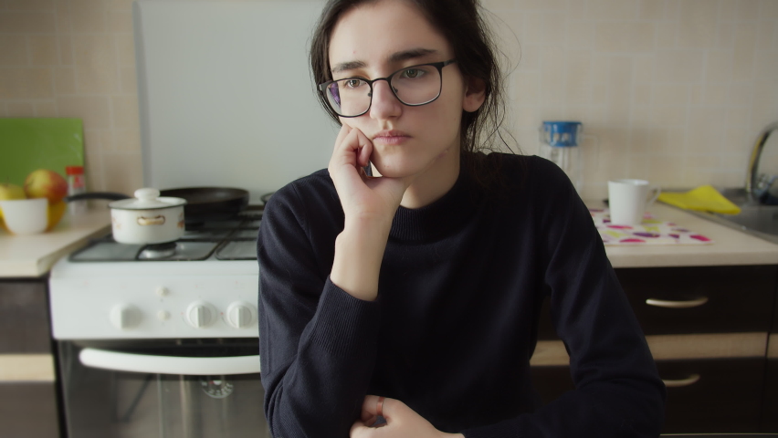 Bored girl is sitting in the kitchen with glasses and thinking about something | Shutterstock HD Video #1061037322