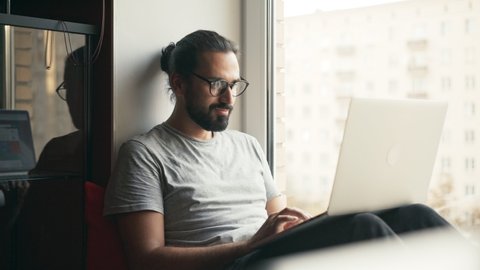 Handheld shot of a young handsome man in glasses working on his laptop while sitting on a windowsill with a city view. Freelancer working from home.