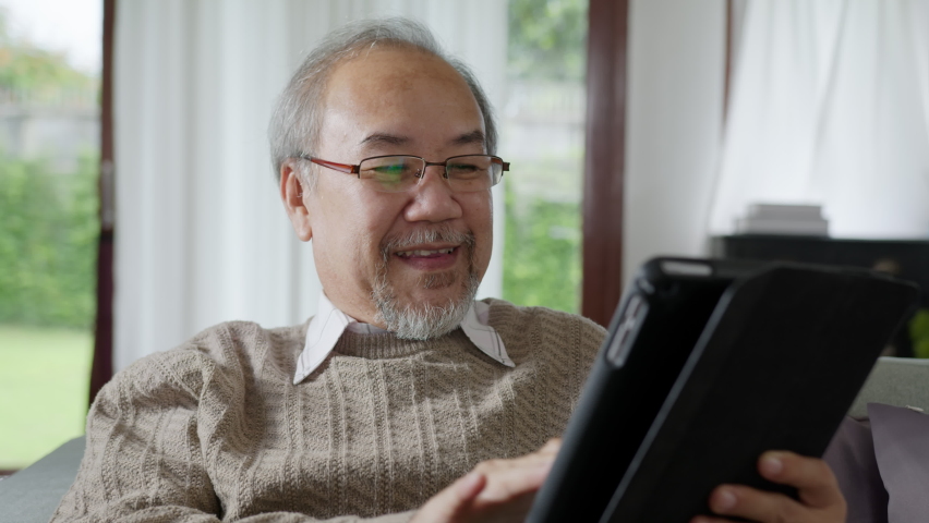 Candid of old asian retired man using tablet computer to transfer money online or financial payment and read text, smile or laugh with happy and positive. Senior asian with digital technology concept