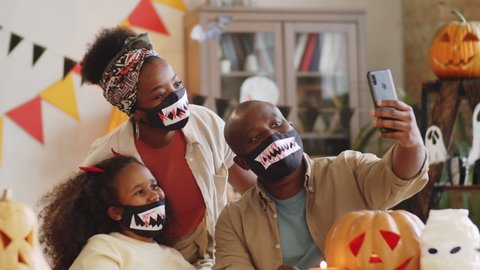 Positive African American family in Halloween face masks with vampire teeth posing and waving at smartphone camera while taking a selfie at home