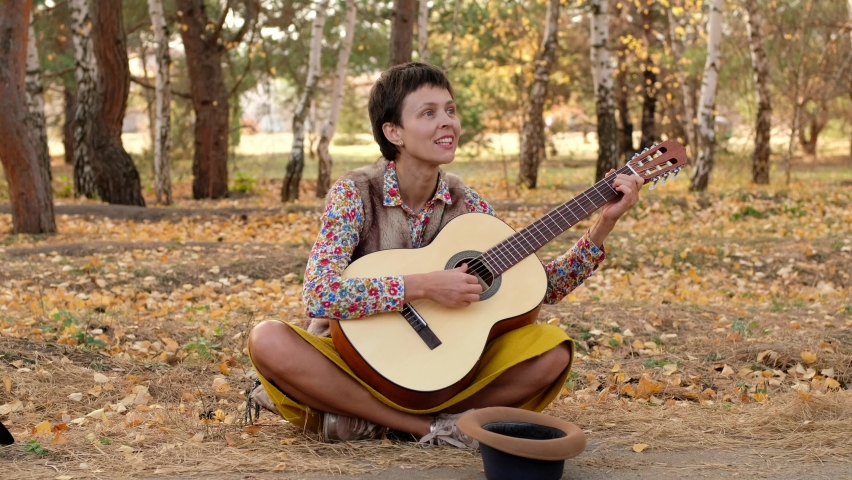 Beautiful woman playing acoustic guitar in park. Street musician performer sitting on ground performs a song asking for money.