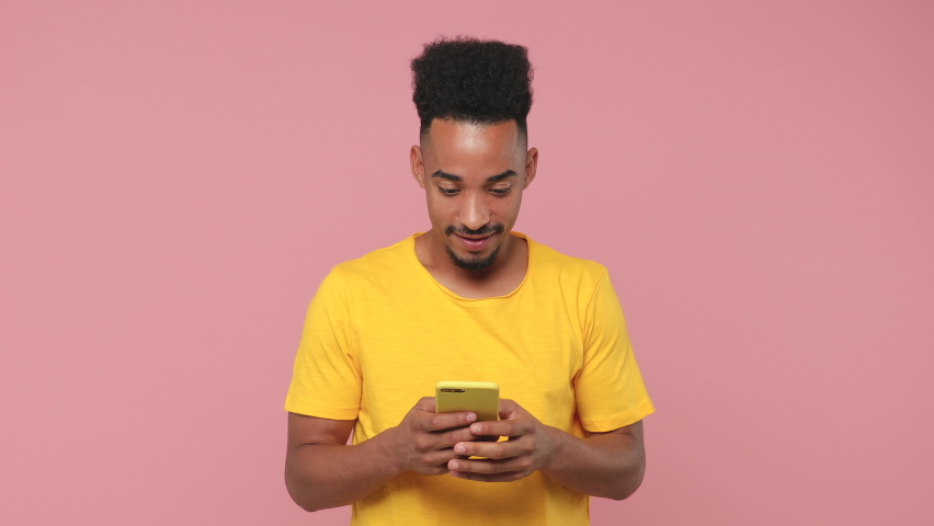 Surprised young african american man in yellow t-shirt posing isolated on pink background studio. People lifestyle concept. Using point finger on mobile cell phone typing sms message showing thumb up