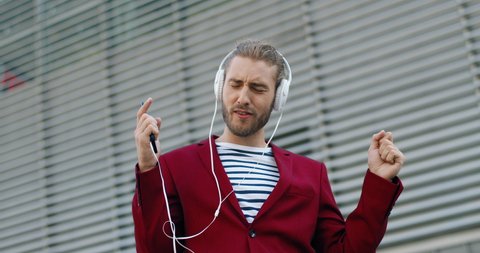 Caucasian young stylish handsome man in headphones listening to music on smartphone and singing. Outdoor. Cheerful good-looking funny guy in red jacket sing and listen to song. Mobile phone player.