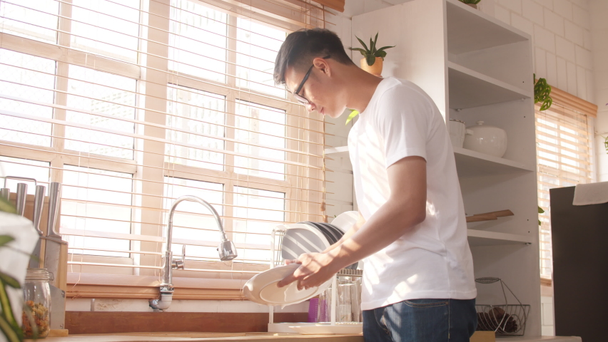 Happy young Asia guy washing dishes while doing cleaning in the kitchen at house. Stay at home, self isolation, social distancing, quarantine for coronavirus, lifestyle man at home concept. | Shutterstock HD Video #1061053195
