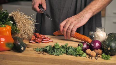 The man is cutting the sausage on the board between the vegetables. Home-made sausage. Everything in the kitchen scenery, the camera slowly moves to the side showing the space of the table.