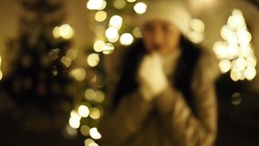 Beautiful girl warming up by illuminated Christmas tree in SLOW MOTION HD VIDEO. Glowing blurred lights decoration in the background. Close-up. Half Speed.