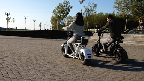 Man and girl on electric scooters are riding through urban seafront at morning. Happy couple is spending time while riding e-bikes next to each other. Ecological electro transport concept.