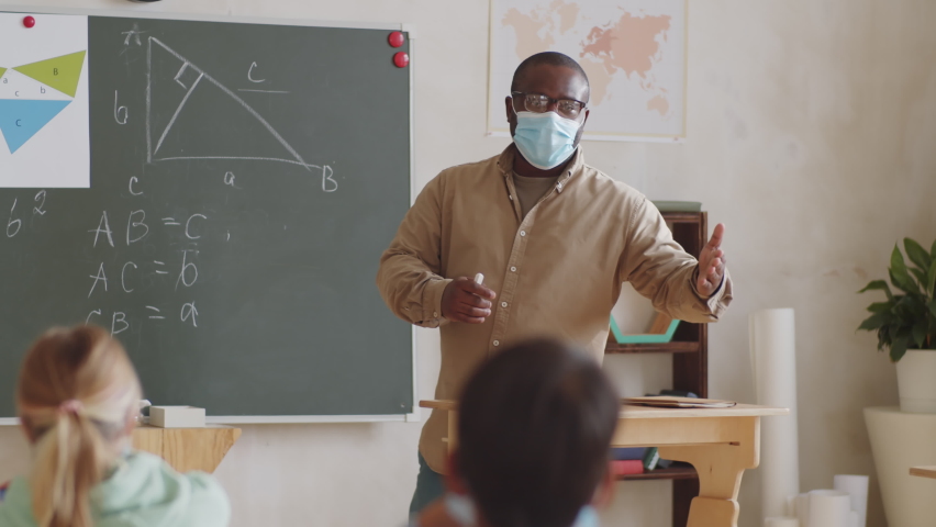 A male teacher in disposable face mask pointing at the chalkboard and giving math lesson to students while working in school during covid-19 pandemic. Royalty-Free Stock Footage #1061063830