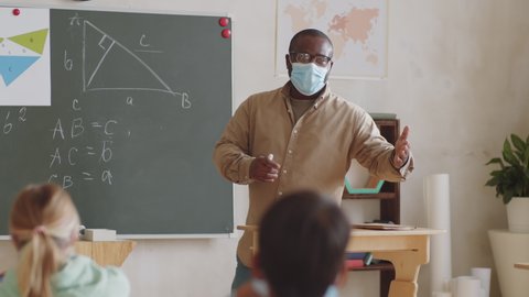 A male teacher in disposable face mask pointing at the chalkboard and giving math lesson to students while working in school during covid-19 pandemic.