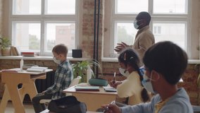 Afro-American male teacher in medical face mask walking through classroom and then standing by lectern and speaking while giving lesson to schoolchildren during coronavirus pandemic. - Powered by Shutterstock - Get 15% off with code: PIKWIZARD15