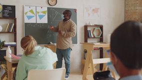 A male teacher in protective face mask standing by chalkboard in front of children in the classroom. pointing at posters and explaining math lesson while working in school during covid-19 outbreak. - Powered by Shutterstock - Get 15% off with code: PIKWIZARD15