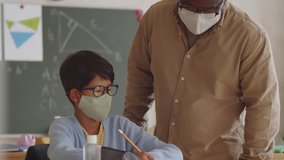 Afro-American male teacher in medical face mask walking through the classroom and helping little students during lesson while working in elementary school during covid-19 outbreak. - Powered by Shutterstock - Get 15% off with code: PIKWIZARD15