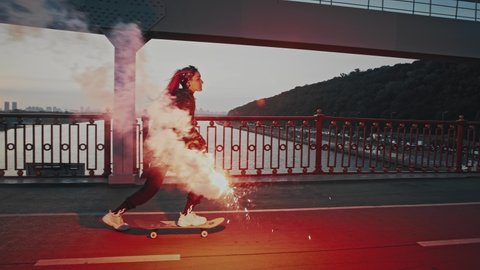 Young hipster female with pink hair, in informal outfit is riding skateboard on bridge holding burning red signal flare and waving it. Riverscape, early morning. Tracking shot. Slow motion, side view
