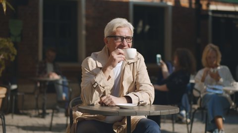 Smiling senior man drinking coffee and flirting with woman sitting at table in outdoor cafe. Portrait of elegant aged man in glasses enjoying coffee and talking to person at next table on terrace