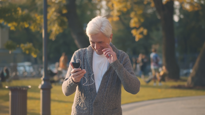 Happy senior man in headphones talking on phone walking in autumn park. Portrait of handsome retired male having video call using earphones and smartphone enjoying walk in city park