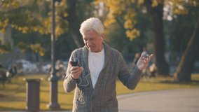 Happy senior man in headphones talking on phone walking in autumn park. Portrait of handsome retired male having video call using earphones and smartphone enjoying walk in city park - Powered by Shutterstock - Get 15% off with code: PIKWIZARD15
