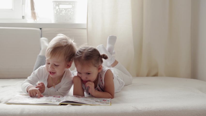 Friendship, family, education concepts  - Two smiling preshool, toddler children read large interesting book of fairy tales on bed. Siblings little twins have fun, happy kids on quarantine at home | Shutterstock HD Video #1061080513