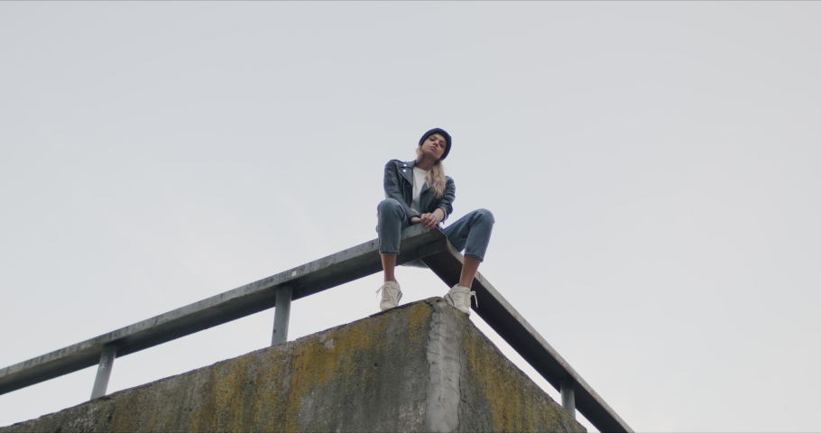 Stylish woman wearing black beanie and leather jacket. Girl siting on rooftop of city building looking at camera. 4k raw slow motion 50 fps