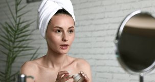 Happy young woman wrapped in bath towel putting cream on face, smiling, looking at mirror and winking to herself. Concept of skincare routine.