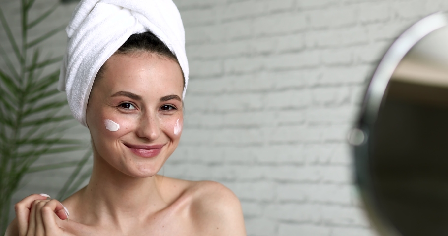 Happy young woman wrapped in bath towel putting cream on face, smiling, looking at mirror and winking to herself. Concept of skincare routine. | Shutterstock HD Video #1061082682