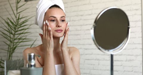 Charming young lady with bare shoulders doing massage on cheeks after morning shower. Happy brunette looking at mirror and doing skin care routine at home.
