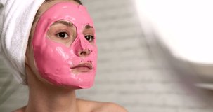 Beautiful young woman with pink alginate mask on face looking at mirror while sitting at bright bathroom. Pretty lady wrapped in bath towel enjoying morning procedures at home.