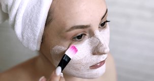 Pleasant young brunette with towel on head using special tool for putting white mask on pure face after morning shower. Concept of skin care routine.