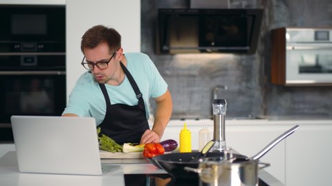 Young man watching video recipe on laptop while cooking vegetables in kitchen. Portrait of handsome guy in apron and glasses learning how to cook watching webinar on computer at home kitchen