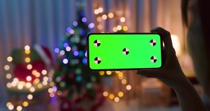 back of asian woman watch video by green screen smartphone with christmas decoration and tree at home