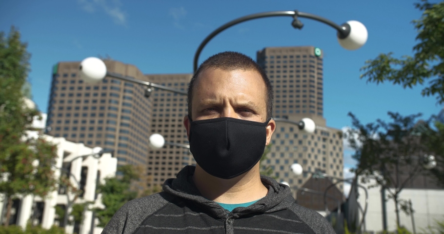 Portrait of handsome young man wearing covid-19 protective face mask and looking upwards with hope standing against buildings in city