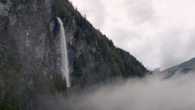 Drone video of a waterfall in the mountains. Flying through clouds and stream of water falls from the rocks.