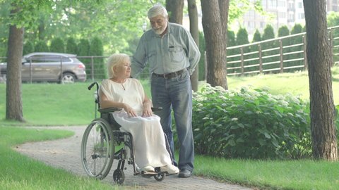 Wide shot of loving old husband giving gift box to paraplegic wife in sunny park. Portrait of affectionate Caucasian man surprising woman. Thankful disabled retiree kissing spouse on cheek.