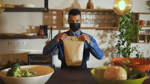 African american waiter wearing protective face mask giving bag to customer at counter bar. Young man worker working with take away orders during corona virus outbreak. Concept food & drinks takeaway