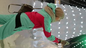 Girl in snowboard clothes holds a green sparkler in her hands on a blurred background of an ice Christmas tree with glowing twinkling garlands. Vertical Video