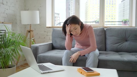 Depressed asian ethnicity young woman sitting on sofa, thinking of personal problems alone at home, unhappy tired mixed race lady suffering from mental stress and feeling sad