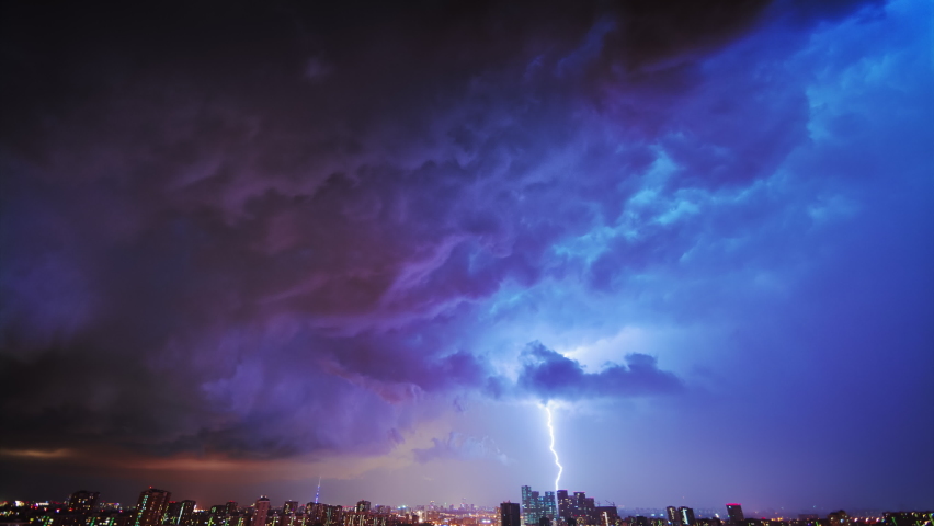 Amazing timelapse of the night storm over the big city, with colorful clouds moving on the sky and bringing first drops of rain, and bright lightnings illuminating the scene.