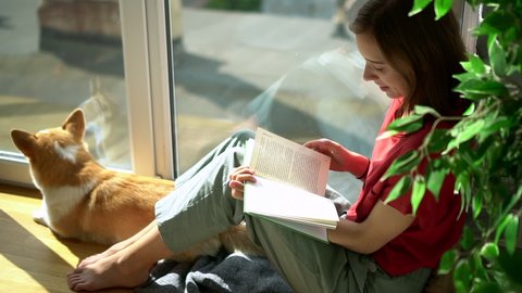 young woman sit by balcony window and read book with cute corgi dog Avki. relaxing indoors near terrace with literature and sleeping pet. sunny footage of female in comfortable windowsill. floor