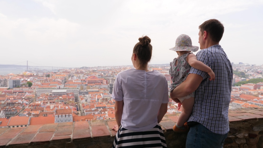 Woman and man enjoy old city panorama, little baby turn round and point to other side by hand, want to explore place. Young tourist family at observation deck. Beautiful Lisbon cityscape on background