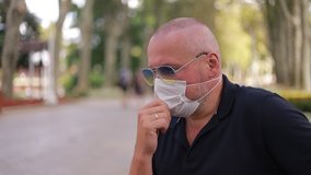 Video, an adult man in a black polo shirt and sunglasses removes and puts on a white protective mask. Outdoors in the park on a sunny day in summer.