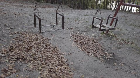 Cloudy autumn day. Fall dark forest, playground in clearing. Dry leaves on ground. Old rusty metal swing swaying in wind. Nobody. Loneliness, loss, depression, lost concept.