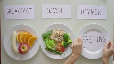 Hands of a woman put a plate with cut fresh fruits and brown rice with vegetables under the titles breakfast and lunch respectively, after which under the title dinner she puts empty plate with title