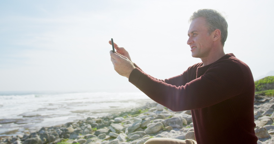 Caucasian man enjoying free time by the sea on a sunny day sitting and using smartphone, in slow motion. Early retirement by the coast.