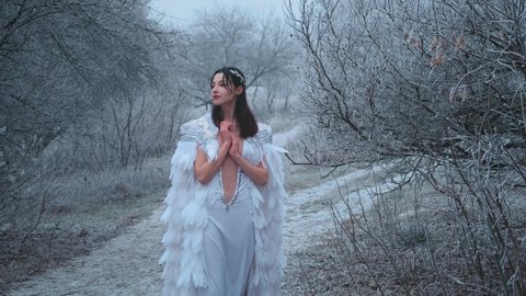 Mystical fantasy Greek goddess walks in snowy forest. Gently touches frosty tree branches. Fairytale princess enjoy winter nature. White vintage carnival art wedding creative cloak cape, bird feathers