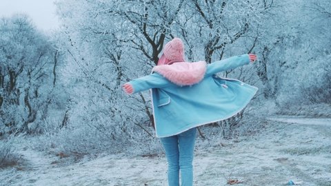 Joyful cheerful happy young beautiful woman runs in winter frosty snowy forest. Girl fun dancing spinning. Pink long loose hair fluttering wind. Warm knitted hat sweater blue denim jacket fox fur hood