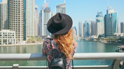 Young happy redhaired woman tourist stands on bridge. Promenade Arab city Dubai Marina. Blue white facade modern high-rise buildings skyscrapers harmony with color sky, water, backpack, hat. UAE 4k 