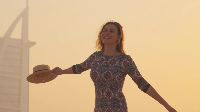 happy cheerful beautiful woman tourist. Girl laughs has fun dancing in Dubai city beach. Hands raised to yellow sky sunset. blond long hair flying in motion. Straw beach hat. UAE 2020 4k Go Everywhere Royalty-Free Stock Footage #1061120866