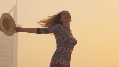 Young happy cheerful beautiful woman tourist. Adult Girl laughs has fun dancing in Dubai city beach. Hands raised to yellow sky sunset. blond long hair flying in motion. Straw beach hat. UAE 2020 4k