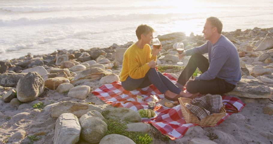 Caucasian couple enjoying free time by the sea on a sunny day drinking wine and talking, in slow motion. Early retirement by the coast.